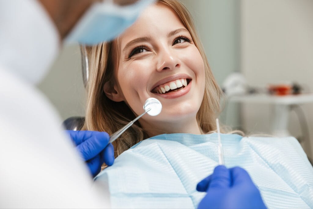 Smiling young woman sitting in a dental chair during a check-up, as a dentist uses dental tools to examine her teeth.