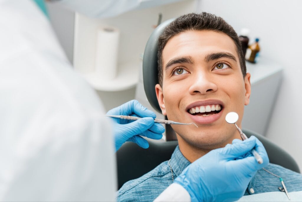 Smiling young man sitting in a dental chair during a dental exam, looking at the dentist while instruments are used to inspect his teeth.