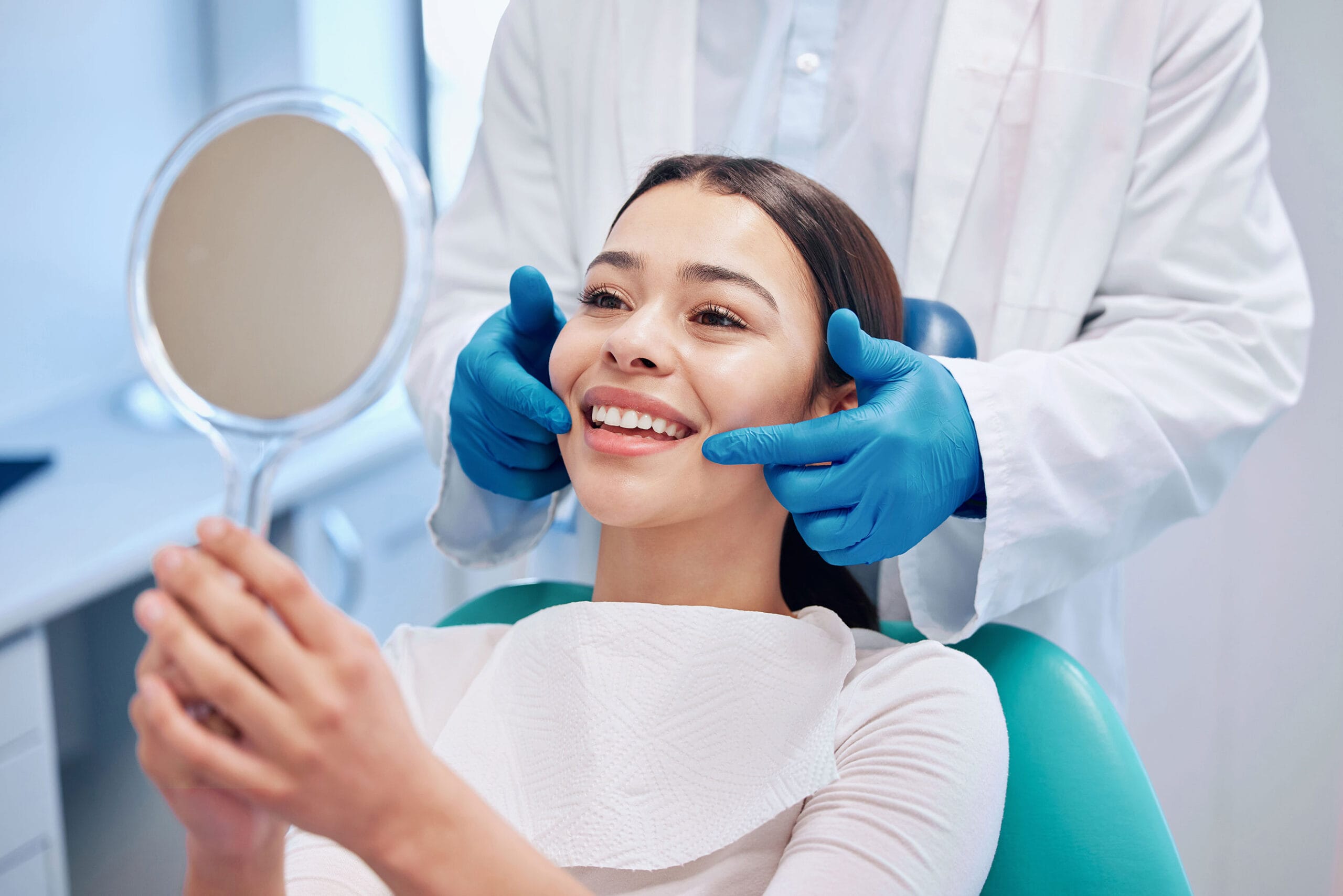 Smiling patient holding a mirror while dentist checks teeth in a bright dental office.