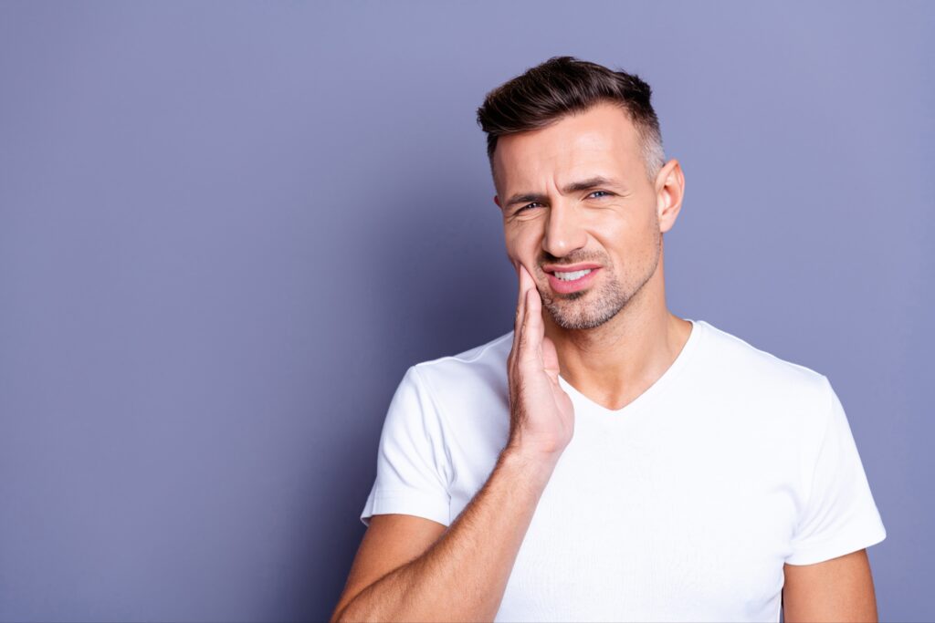 Man in a white t-shirt holding his cheek and wincing in discomfort, indicating dental pain or trauma, against a plain purple background.