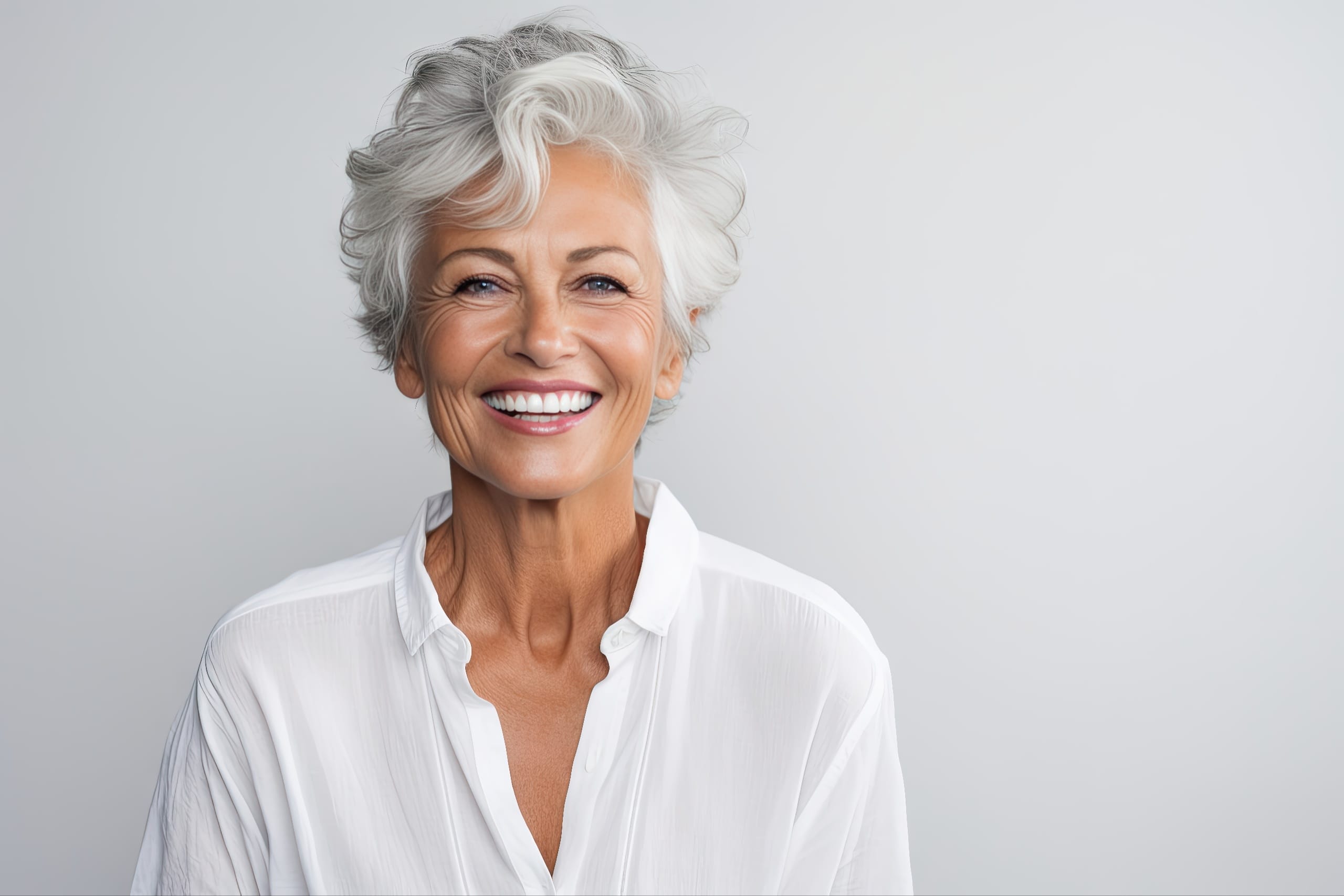 Confident older woman with short white hair smiling brightly, showing perfect teeth while wearing a white blouse against a neutral background—suggesting successful denture treatment.