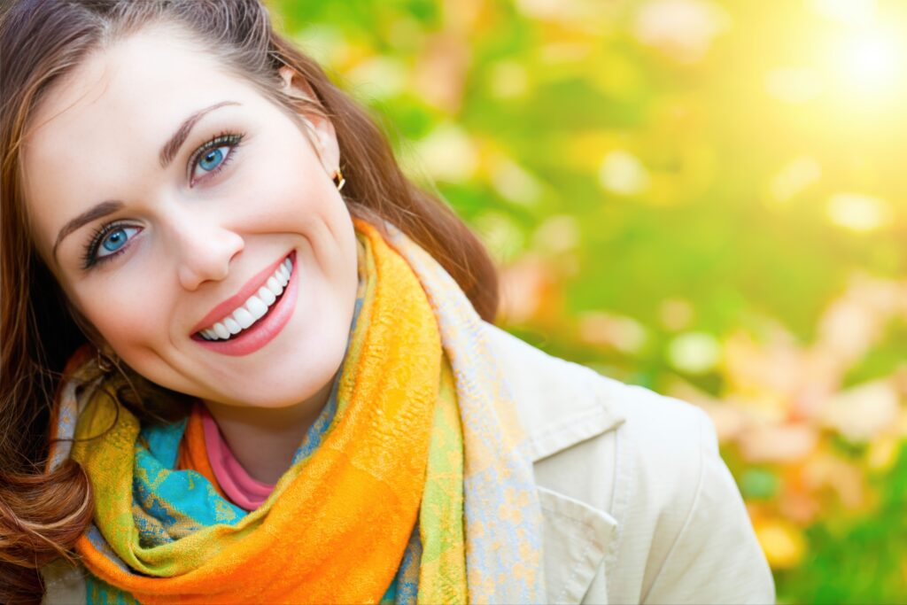 Close-up of a smiling woman with bright white teeth and blue eyes, wearing a colorful scarf and light jacket outdoors with a blurred autumn background and sunlight.