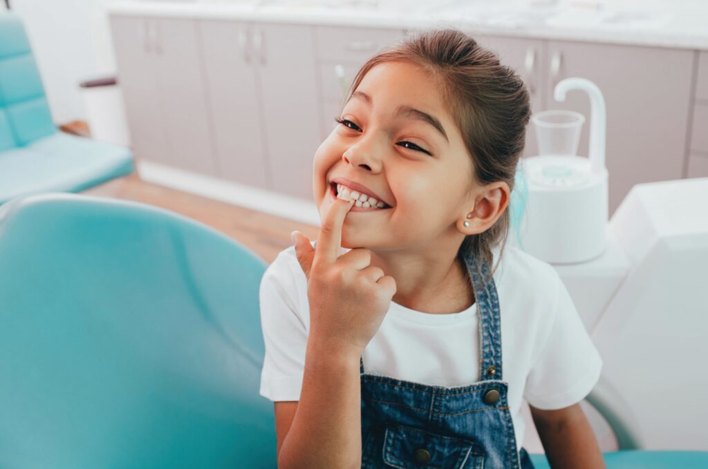 Smiling young girl in a dental clinic pointing to her teeth with her index finger, wearing a white shirt and denim overalls while sitting in a dental chair
