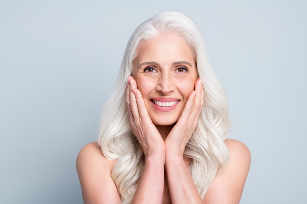 Smiling mature woman with long white hair gently holding her face, showcasing healthy, restored teeth—symbolizing successful restorative dentistry.