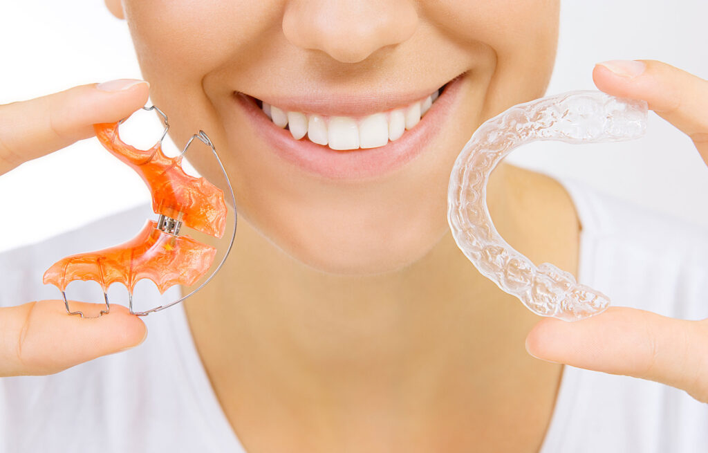 Person smiling while holding a traditional wire retainer in one hand and a clear aligner in the other.