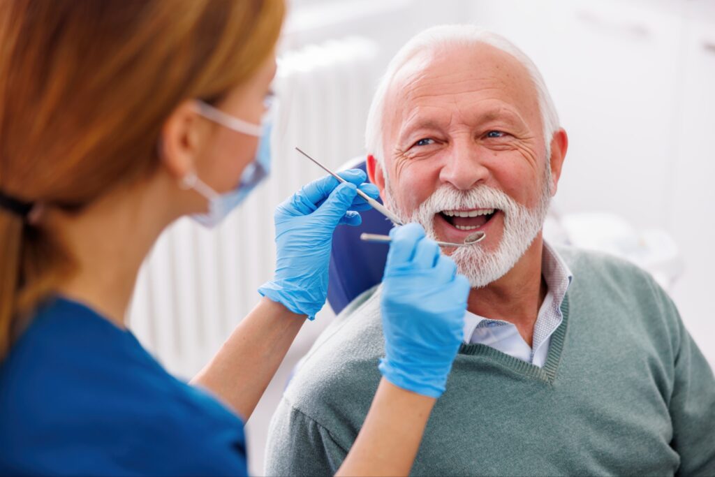 Smiling senior man in a dental chair receiving an oral examination from a dental professional wearing blue gloves and a face mask, representing sinus lift consultation or dental care for older adults.