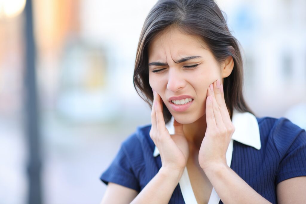 Young woman outdoors holding both sides of her jaw with a pained expression, indicating TMJ (temporomandibular joint) discomfort.