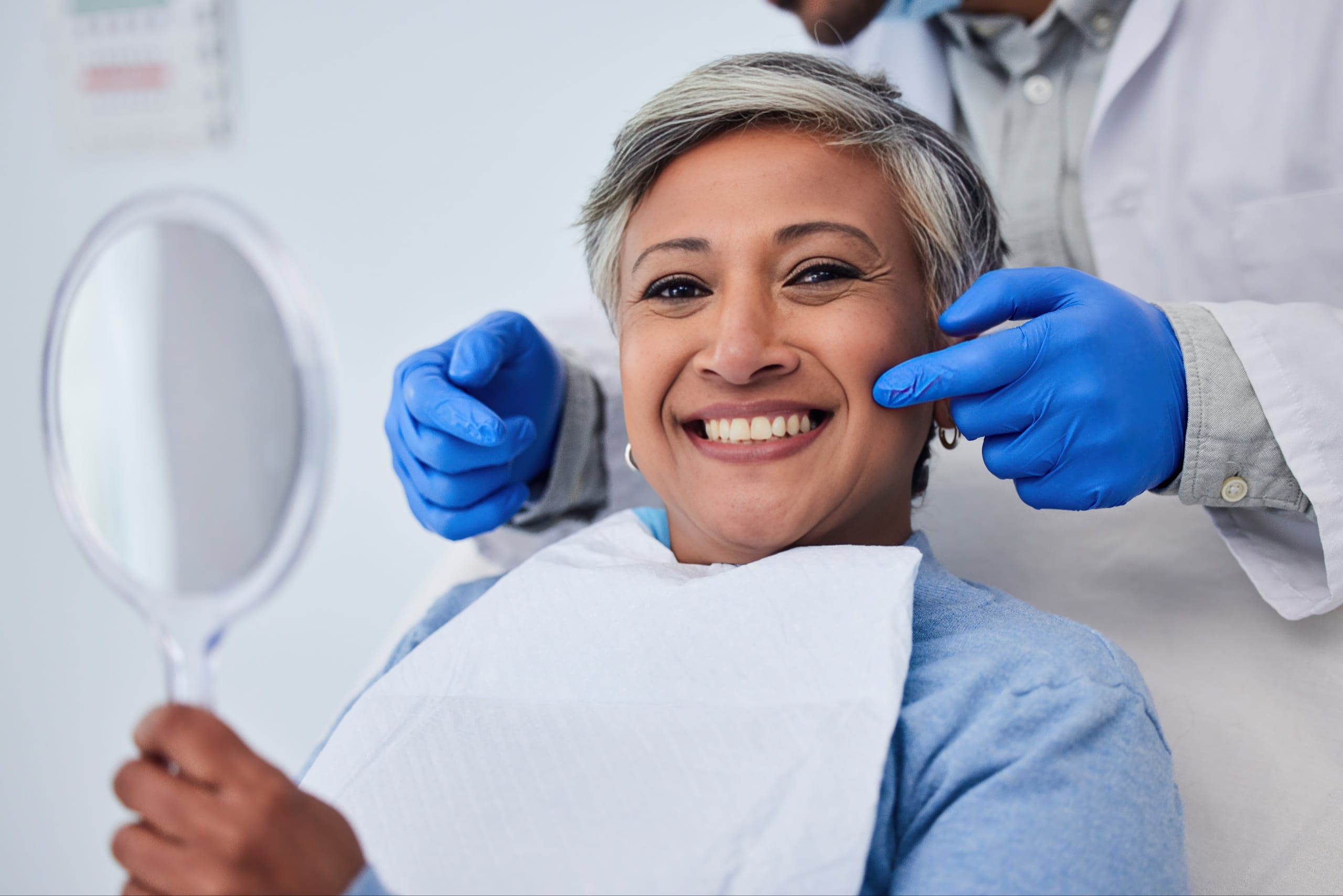 Smiling female patient holding a mirror while the dentist points to her teeth after a successful tooth bonding procedure.