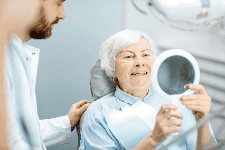 Elderly woman smiling while checking her teeth in a mirror during a dental visit.