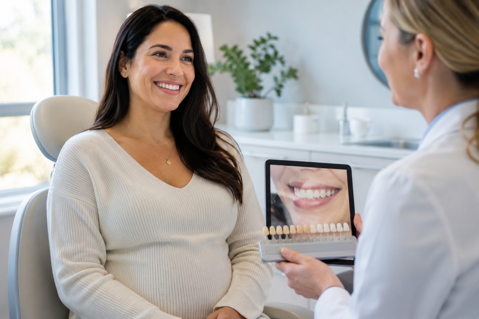 Cosmetic Dentistry Smiling adult seated in a modern dental office during a cosmetic dentistry consultation while a clinician holds a tablet showing a smile design image and a dental shade guide nearby.